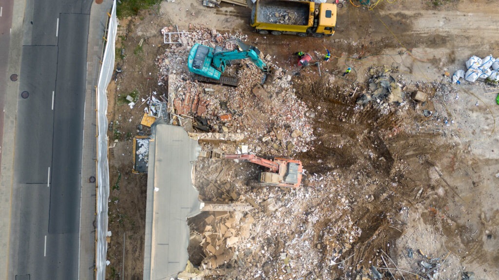 Aerial view of a demolition site with two excavators and a yellow dump truck clearing debris from a demolished building. Rubble and construction materials are scattered around. Two workers are visible among the debris.
