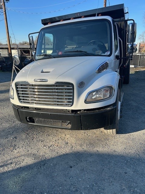 A white Freightliner truck is parked on a gravel lot in daylight, facing the camera. There is a black dump bed attached to the truck and some fencing and utility poles visible in the background.