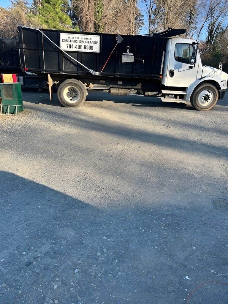 A white dump truck with a black dumpster container is parked on a gravel lot. A sign on the container reads “Dumpster Rental, 704-400-6990.” Trees and clear blue sky are visible in the background.
