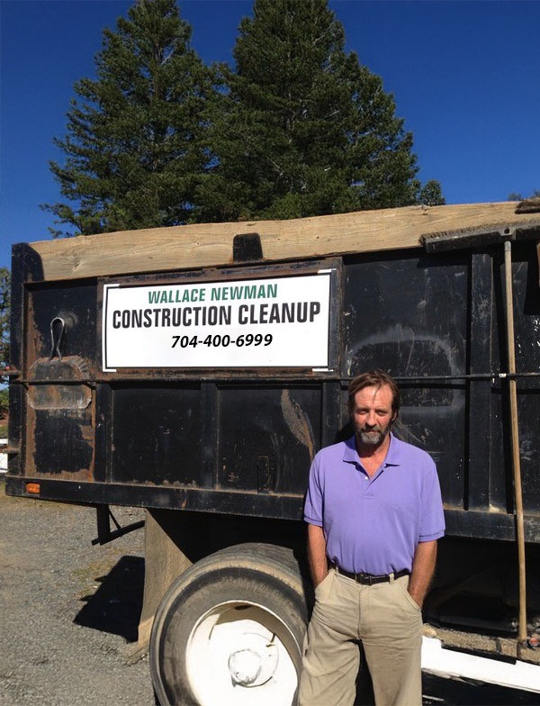 A man in a light purple polo shirt stands in front of a black construction cleanup truck with a "Wallace Newman Construction Cleanup" sign and a phone number on the side, with tall green trees and a clear blue sky in the background.